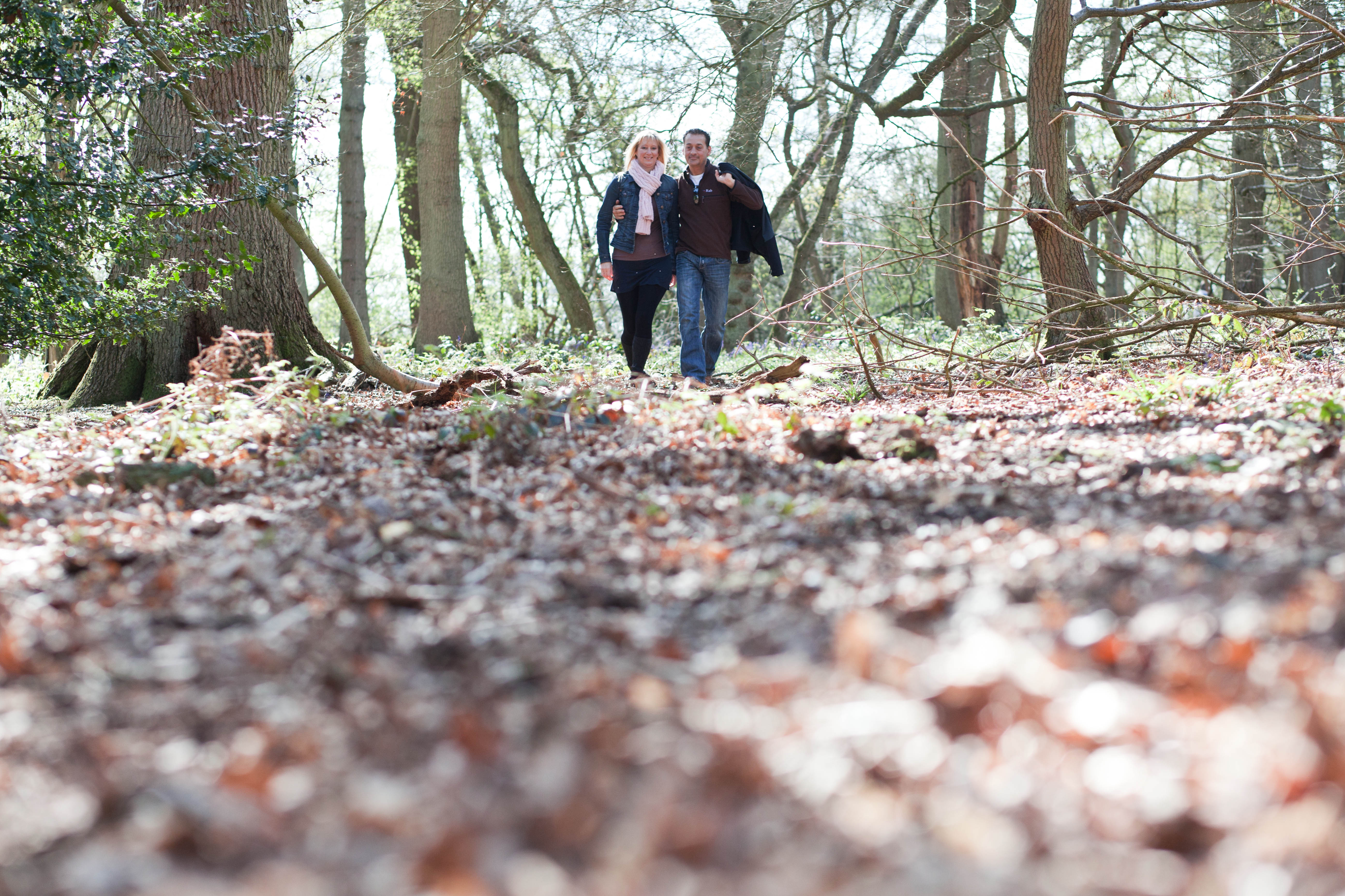 Man and lady arm in arm walking through wood across autumn leaves.