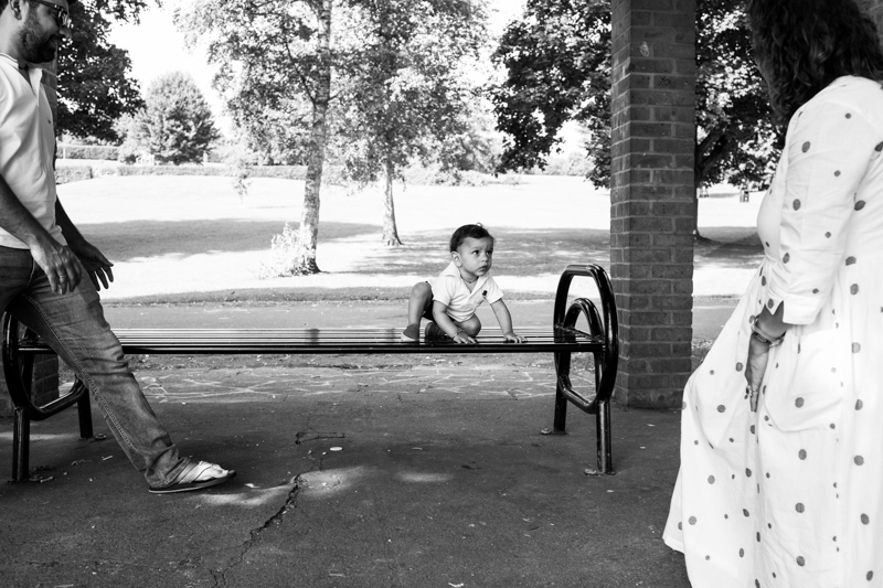 Baby boy on a bench, his parents walking towards him. 