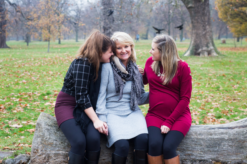Two ladies cuddling their mum, while they sit on a tree trunk in the park