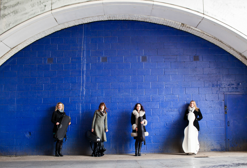 Four ladies with musical instrument cases in front of huge blue wall. 