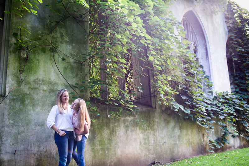 Mother and daughter standing in front of derelict church wall covered in greenery. 