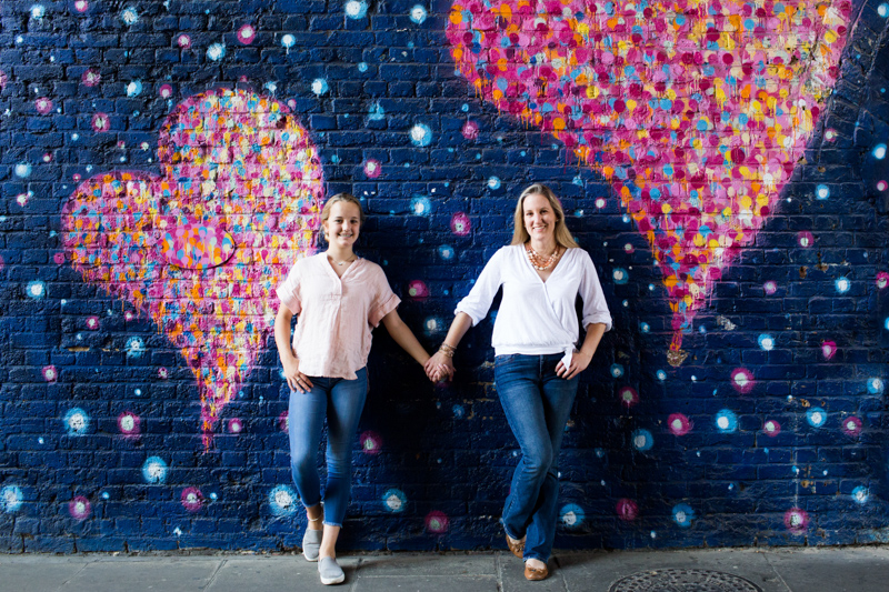Mum and daughter holding hands in front of heart wall. 