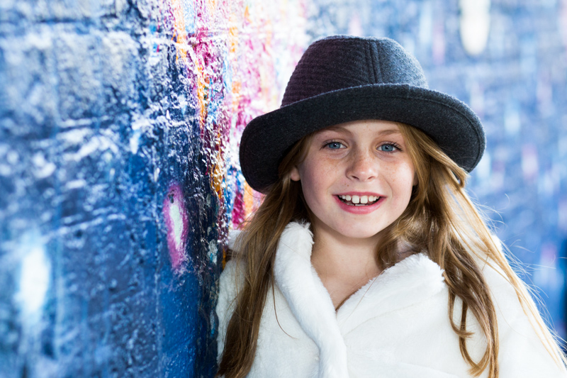 Girl wearing a hat, leaning on colourful wall.
