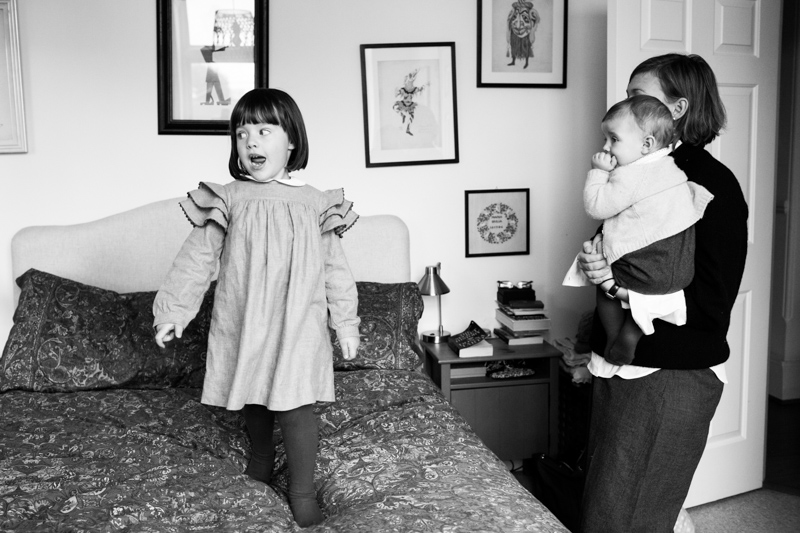 Little girl pulling a funny face on a bed, with mum and baby looking on.