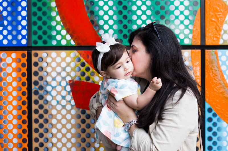 Mum kissing her baby girl in front of colourful wall. 
