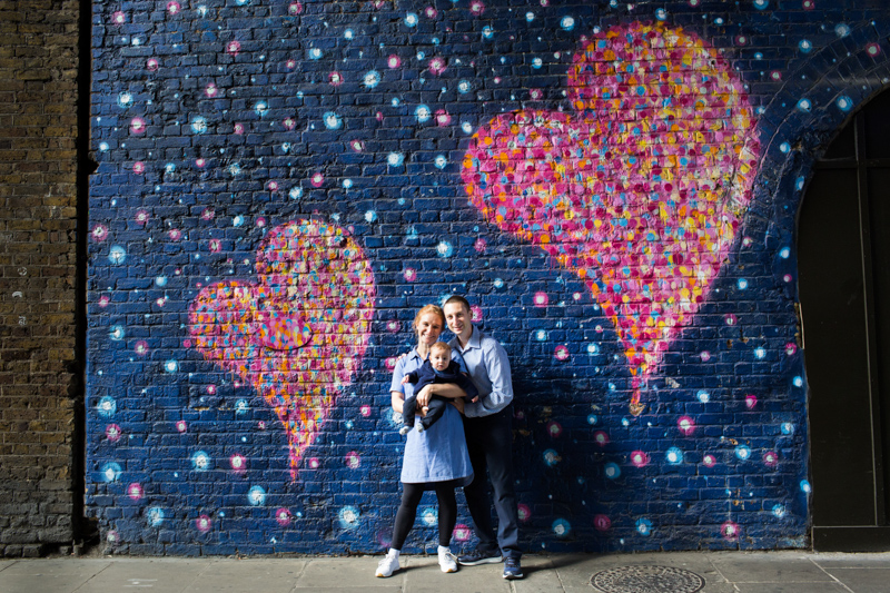 Lady, man and little baby standing between two large hearts on drawn on a brick wall.