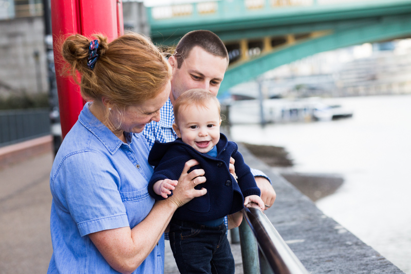 Baby smiling with lady and man, with green bridge in the background. 