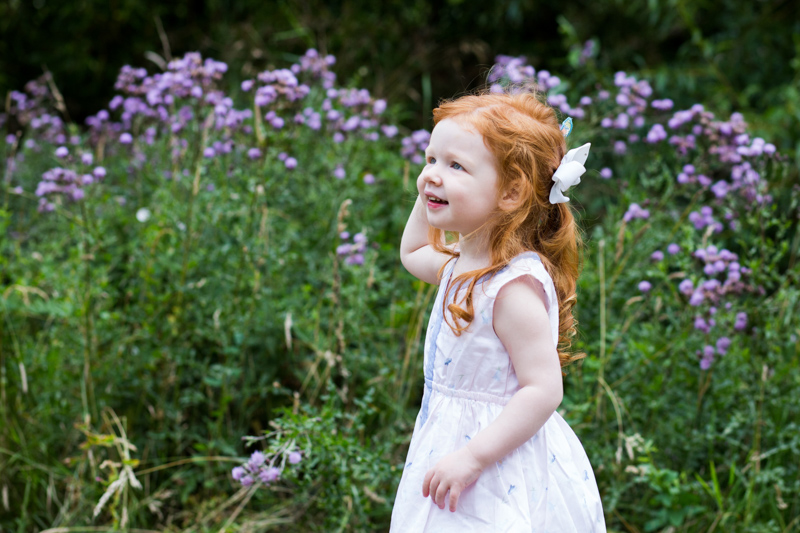 Girl with red hair and white dress in front of purple flowers. 
