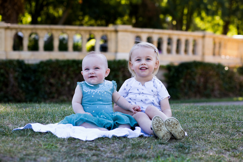 Two sisters sitting on a white blanket on the grass.