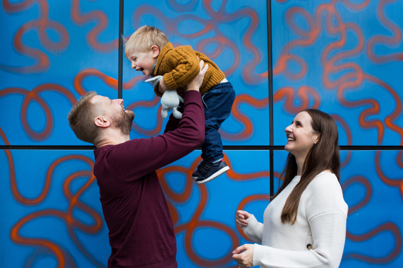Boy lifted up by dad in front of colourful wall. 