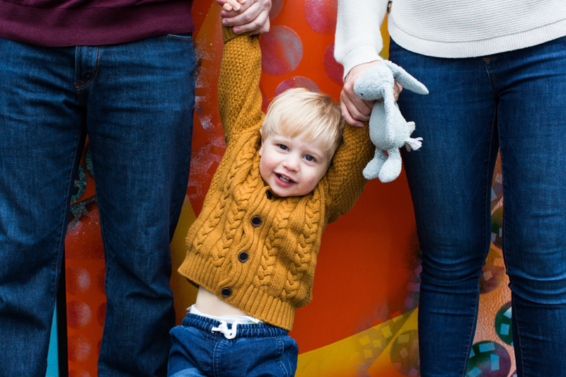 little boy holding mum and dad's hands in front of colourful wall. 