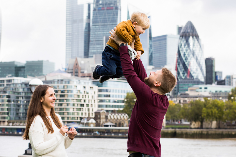 Dad lifting son up high with the City of London in the background and mum looking on. 