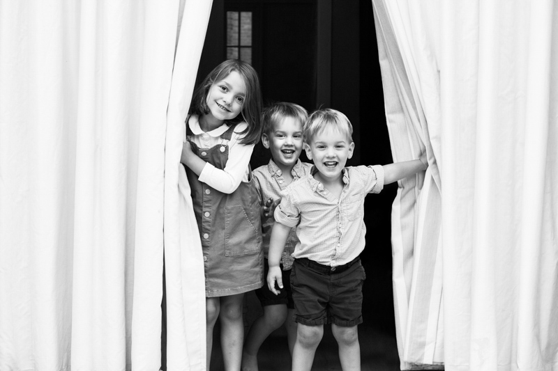 Three children smiling through open curtains.