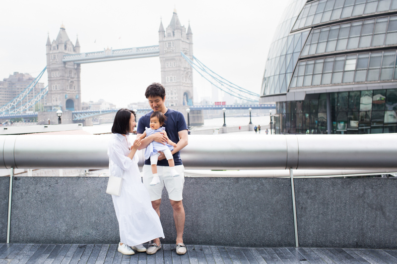 Mum, Dad and baby girl standing in front of the River Thames, Tower Bridge and City Hall in London. 