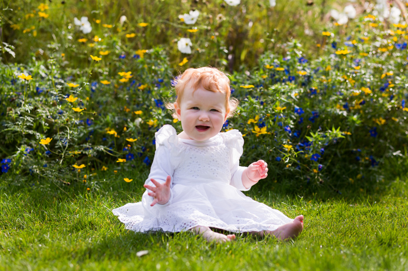 Baby sitting on grass in front of beautiful flowers. 