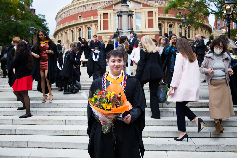 Young man with graduation gown and flowers in front of busy scene. 