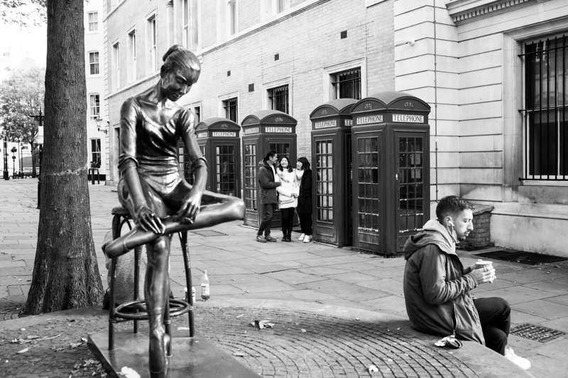Family of three by some phone boxes, with a ballerina statue in the foreground 