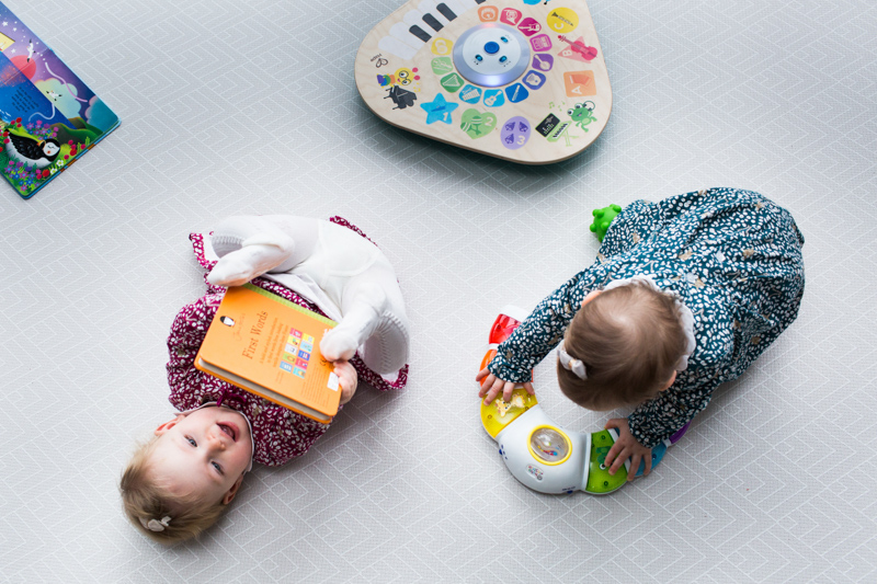 One girl lying on her back with a book, the other girl playing with a toy. 