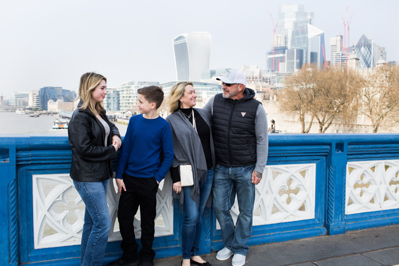 Family of four standing on a blue bridge with the City of London in the background. 