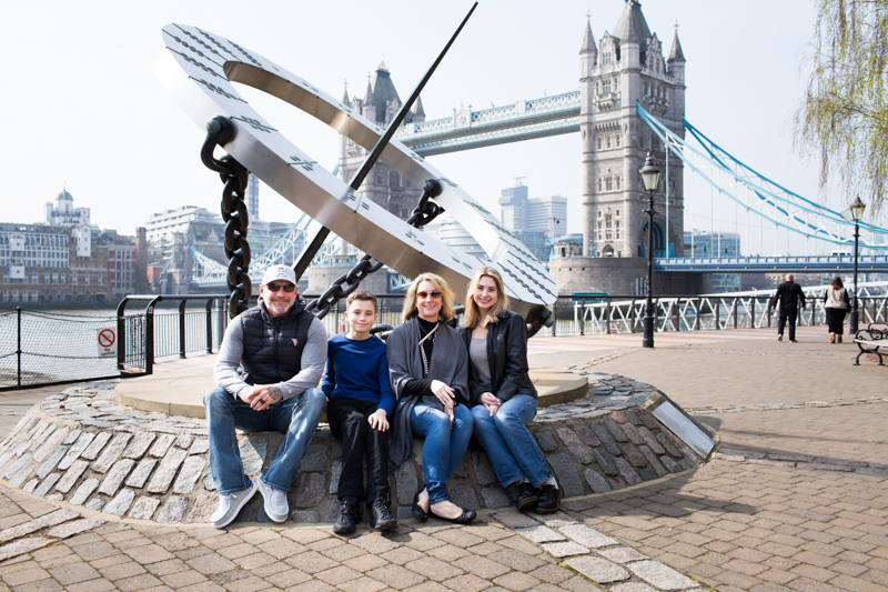 A man, a boy and two ladies sitting down with Tower Bridge in the background. 