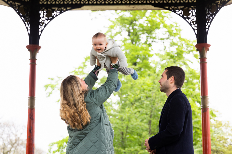 lady holding baby up in the air, while man looks on