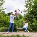 Man holding little girl up in the air, while lady looks on.