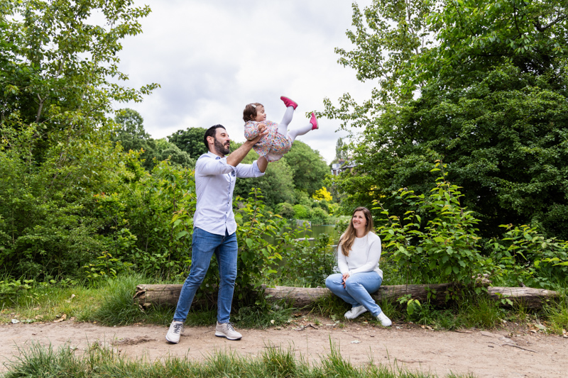 Man holding little girl up in the air, while lady looks on.