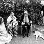 2 older people in chairs, younger woman sitting on the grass, man with hands on hips looking at girl lying on floor