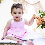 baby girl in pink dress touching some flowers with a "one" sign behind her