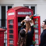 lady holding baby up in front of a london red telephone box, as man watches on smiling
