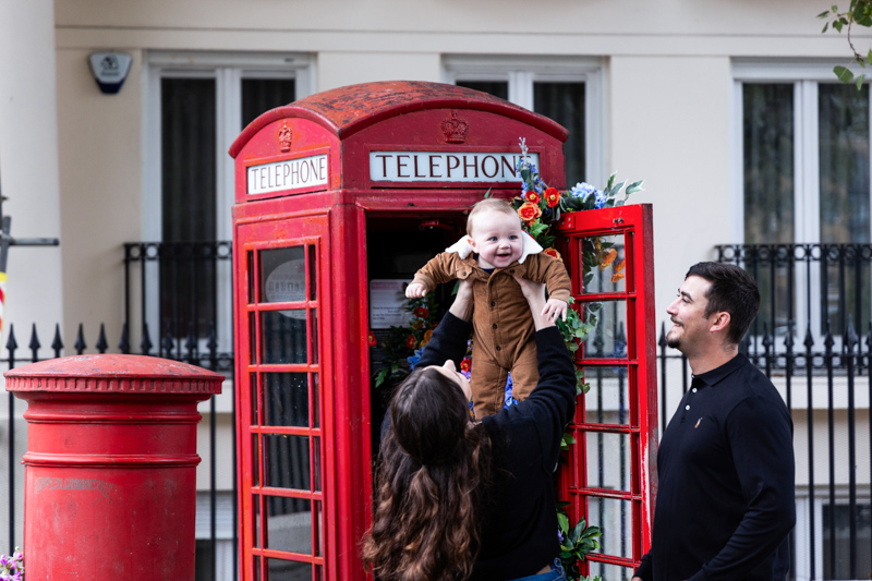 lady holding baby up in front of a london red telephone box, as man watches on smiling