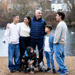 Family of six, and their dog, smiling at each other with a pond behind them.