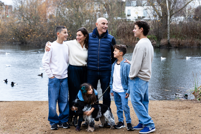 Family of six, and their dog, smiling at each other with a pond behind them.